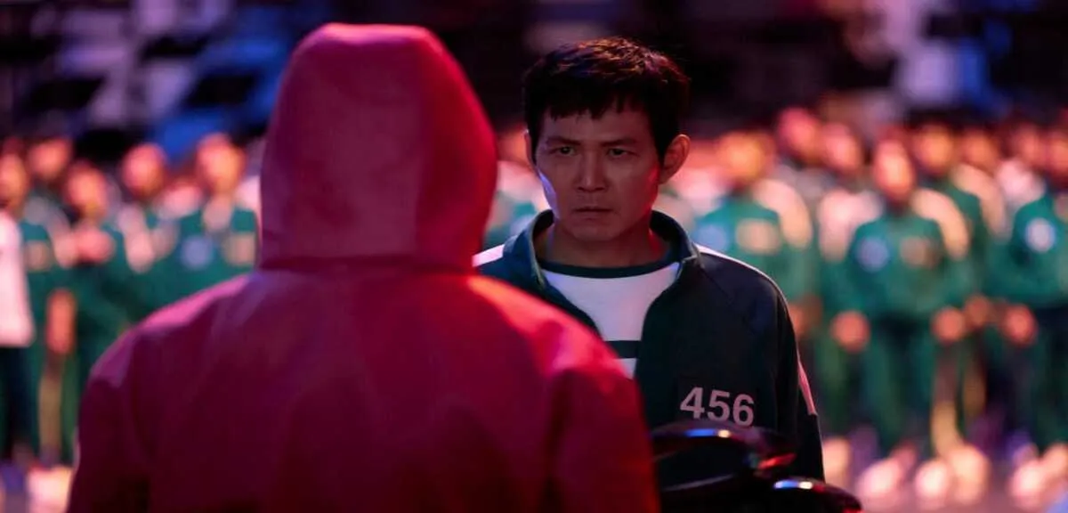 Squid Game dizisinin gergin bir anında, 456 numaralı oyuncu Seong Gi-hun'un kararlı ve gergin yüz ifadesi, ön plandaki kırmızı kapüşonlu bir figürün arkasından net bir şekilde görülürken, arkada bulanık yeşil eşofmanlı diğer oyuncularla çevrili olduğu dramatik sahne.