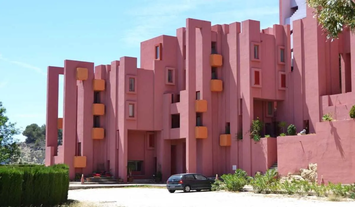 Parlak güneş ışığı altında, keskin geometrik hatlara ve pembe tonlara sahip La Muralla Roja konut kompleksi; renkli, labirent benzeri mimarisiyle Squid Game dizisinin merdiven setlerini görsel olarak çağrıştırıyor.