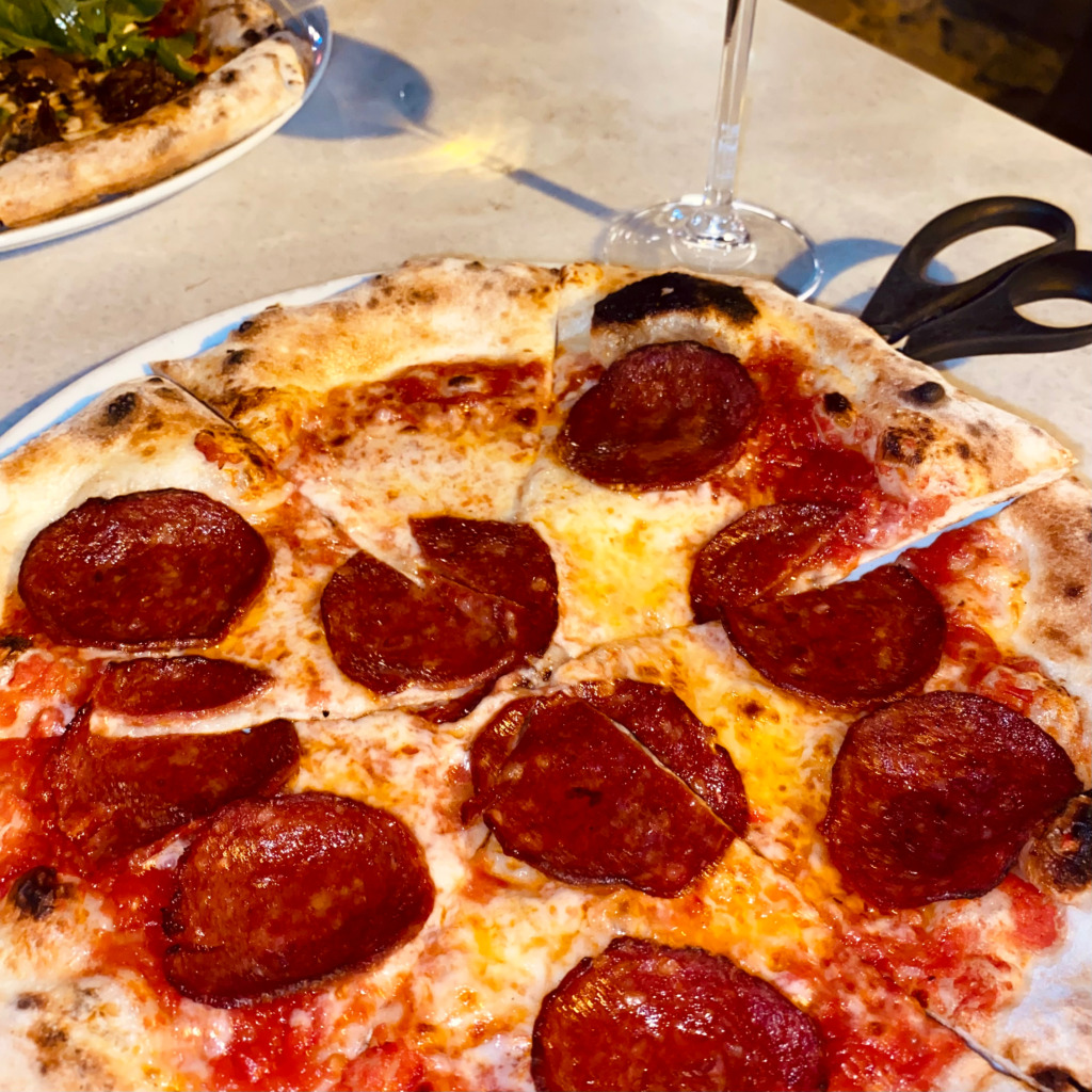 Cunda'nın kendine özgü atmosferinde, adanın gastronomik lezzetlerini tamamlayan, ince ve çıtır hamurlu bol sucuklu bir pizzanın yanında şarap kadehi ve pizza makasıyla sunulduğu keyifli bir akşam yemeği anı yakalanmış.