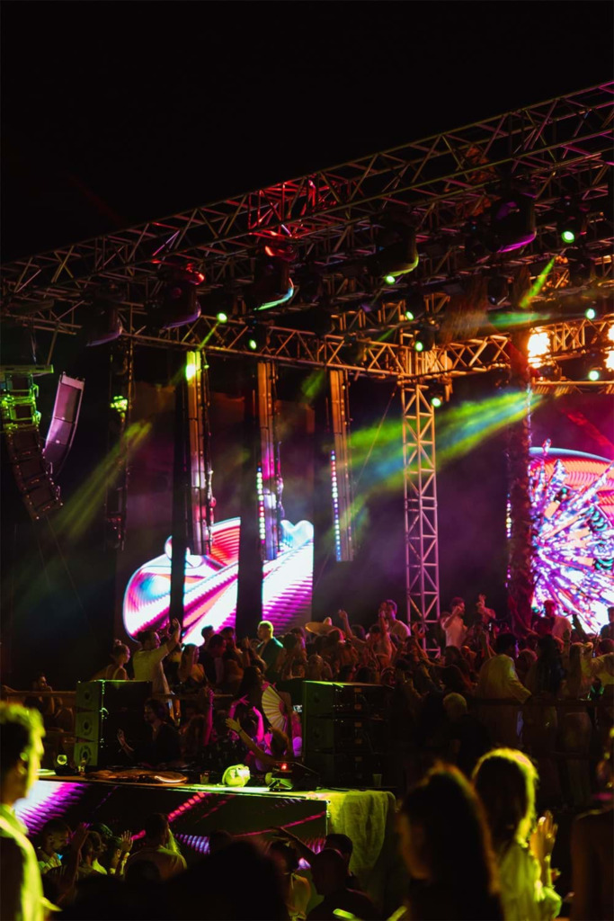 Çeşme'nin en gözde gece mekanlarından birinde, metal iskelelerle çevrili ve rengarenk ışıklarla aydınlatılan sahnede çalan DJ'in ritmine eşlik eden, ellerini havaya kaldırmış coşkulu kalabalık dans ederken, LED ekranlardaki hareketli görseller atmosferi daha da yükseltiyor.