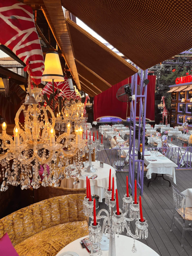 Çeşme'nin hareketli gece mekanlarından birinde, parıldayan kristal avizeler, kırmızı mumlu şamdanlar ve sahne arkasındaki kadife perdelerle donatılmış, hem happy hour hem de akşam eğlenceleri için hazır şık bir ortam resmediliyor.