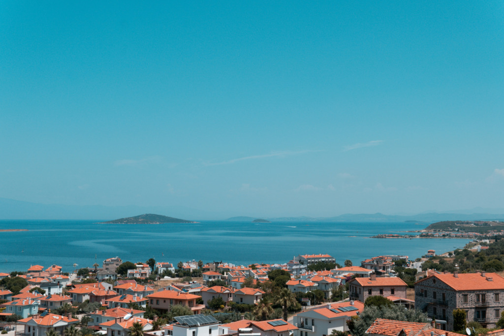 Cunda Seyahat Rehberi başlığındaki adanın karakteristik dokusunu yansıtan bu geniş açılı görüntüde, berrak turkuaz Ege sularının kucakladığı kıyı boyunca sıralanmış, kırmızı kiremitli çatılı taş evler ve yemyeşil ağaçlar, uzakta ise Ayvalık Körfezi'nin dingin adacıkları seçiliyor.