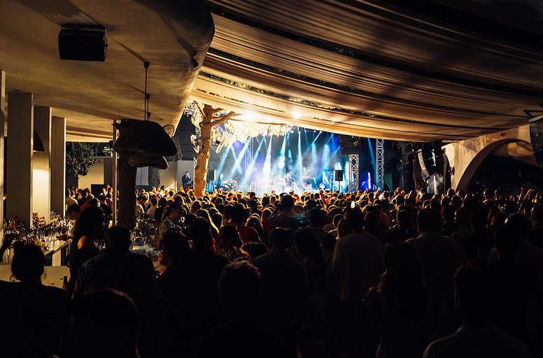 Çeşme'nin en iyi gece mekanlarından birinde, sahnedeki mavi ve beyaz ışıklarla aydınlatılmış canlı müzik performansı eşliğinde dans eden kalabalık, happy hour'dan geceye uzanan enerjik anları yansıtıyor.