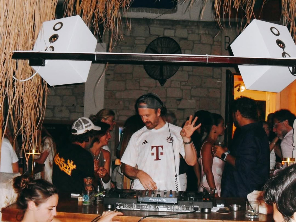 Çeşme'nin en iyi happy hour ve gece mekanlarından birinde, kalabalıklar arasında beyaz tişörtlü bir DJ, Pioneer mikser başında coşkulu bir müzik performansı sergilerken, tavandan sarkan hasır dekorasyon ve modern beyaz hoparlörler eğlenceye ritim katıyor.