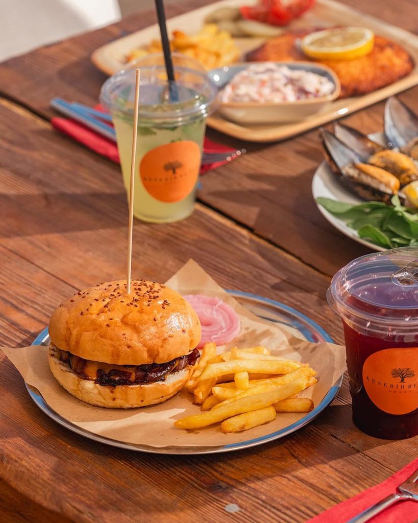 Cunda'da, ahşap bir masada servis edilen lezzetli bir hamburger, patates kızartması ve tazeleyici içecekler, Kesebir Beach'in şık ve doğal atmosferinde bir Cunda seyahat rehberi deneyimini sunuyor.