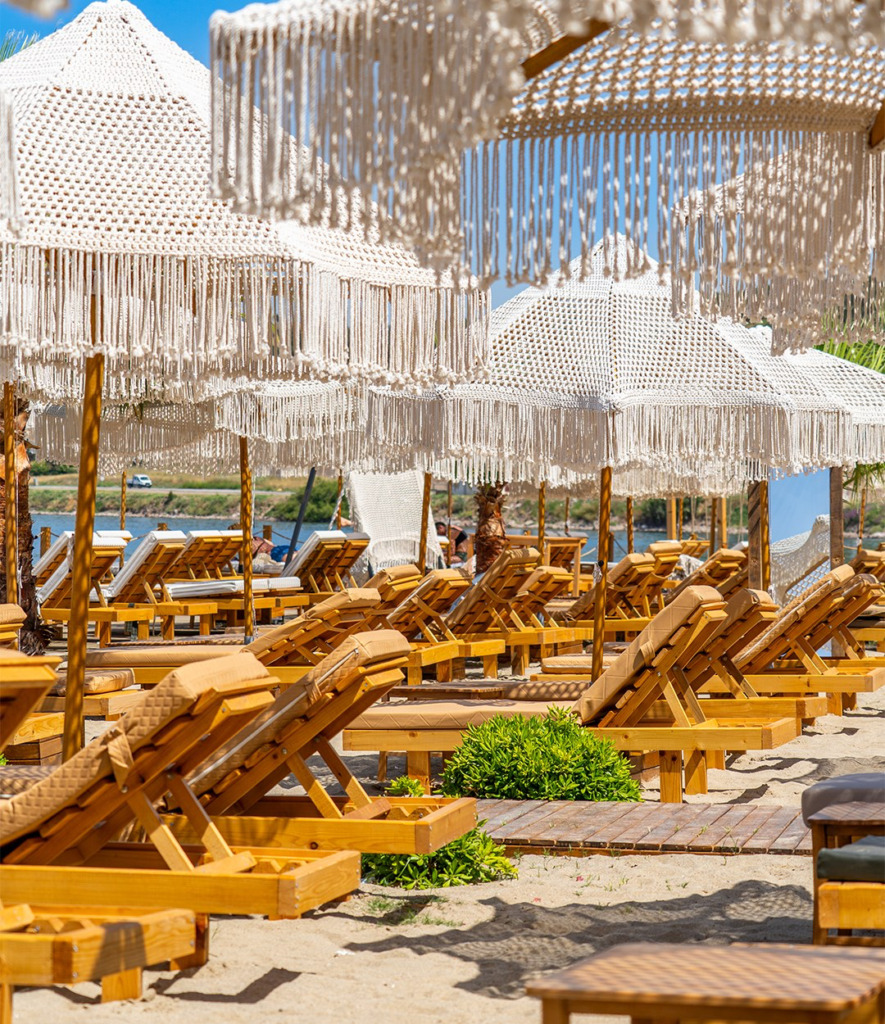 Mavi gökyüzü altında, Cunda'nın sakin plajlarından birinde sıralanmış, boho tarzı püsküllü makrome şemsiyeler ve konforlu ahşap şezlonglar, Ege'nin dingin sularına nazır keyifli bir dinlenme alanı sunuyor.