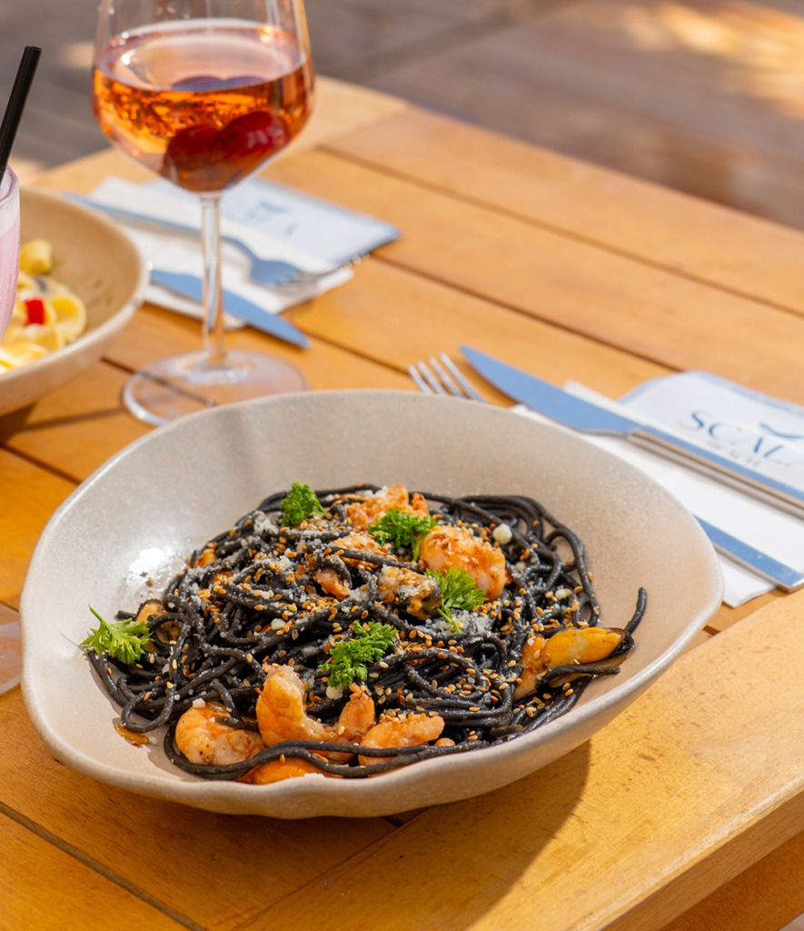 Cunda'da güneşli bir öğle vaktinde Scala Beach'te servis edilen, bol karides, midye, susam ve maydanozla zenginleştirilmiş siyah mürekkep balıklı spagetti tabağı, yanında serinletici bir rose şarap ve yaz kokteyliyle Ege'nin lezzetlerini sunuyor.