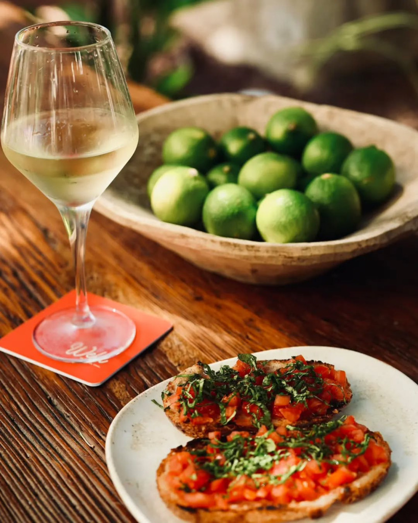 Çeşme'nin en iyi happy hour mekanlarından biri olan Well'de, ahşap bir masanın üzerinde, taze domatesli bruschetta tabağı, serinletici bir kadeh beyaz şarap ve yeşil misket limonlarıyla dolu rustik kase, keyifli bir başlangıcı gözler önüne seriyor.