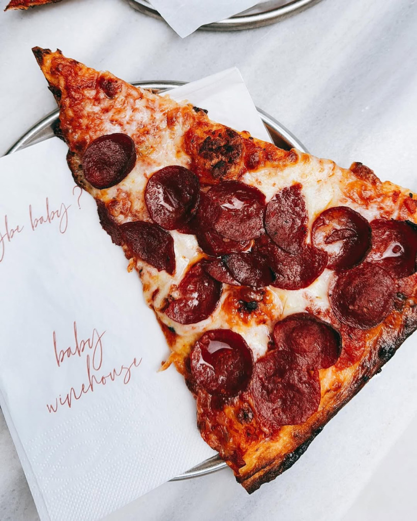 Bir metal tabak üzerinde beyaz bir peçete ve peçetenin üstünde bol peynirli ve bol sosisli, kenarları hafifçe kızarmış bir dilim pepperoni pizza görülüyor, peçetede kırmızı renkle Baby Winehouse yazısı okunuyor ve bu detay Çeşme'nin en iyi happy hour ve gece mekanları arasında yer alan Baby Winehouse'un sunduğu lezzetli atıştırmalık menüsünden bir örneği temsil ediyor.