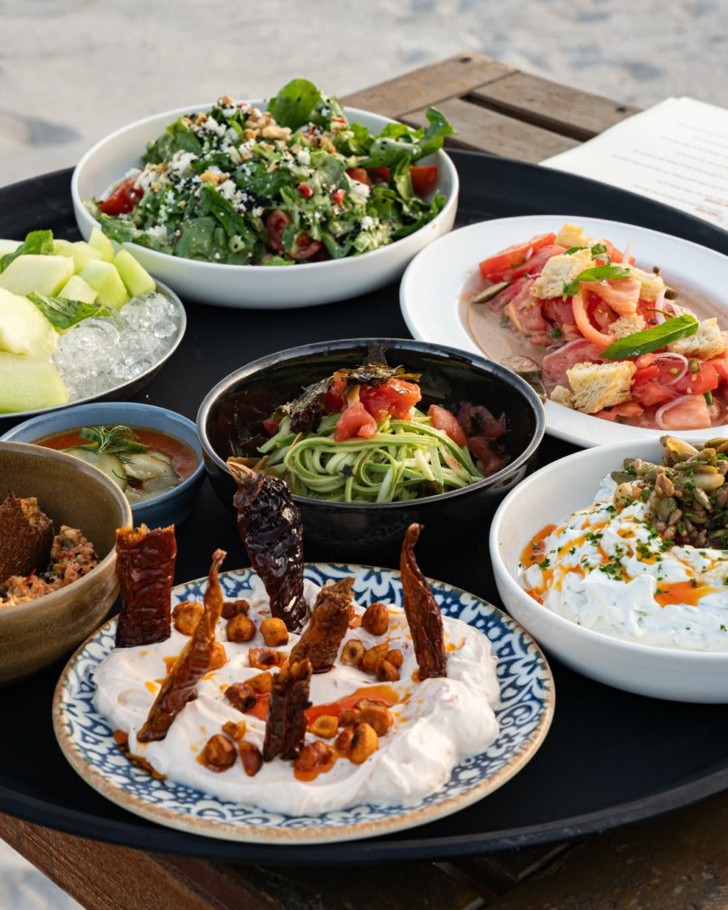 Kumsal kenarında ahşap bir masada sunulan, renkli ve iştah açıcı çeşitli taze başlangıçlar, Çeşme'nin hareketli happy hour'ları ve gece mekanlarında sunulan zengin lezzetleri vurguluyor.