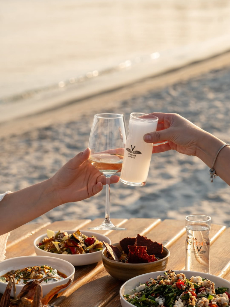 Veranda Alaçatı logosu taşıyan kadehleri ve çeşitli mezeler eşliğinde gün batımında sahilde kadeh tokuşturan iki kişinin mutlu anı, Çeşme'nin en iyi happy hour ve gece mekanları konseptini yansıtıyor.