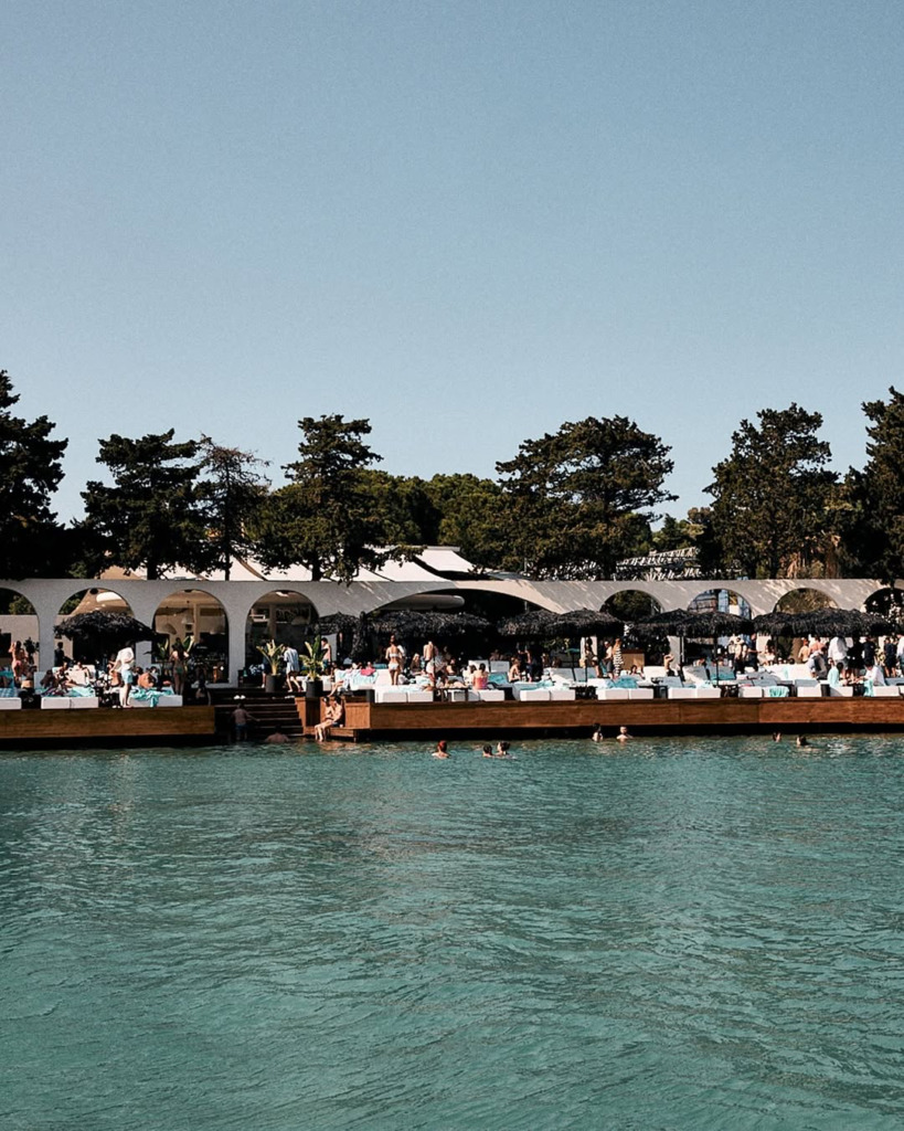 Alaçatı'da deniz kenarında bulunan lüks bir beach club'da, turkuaz renkli suda yüzen ve bembeyaz şezlonglarda dinlenen kalabalık, mekanın enerjik happy hour ve gece eğlencelerini yansıtan canlı bir atmosfer sunuyor.