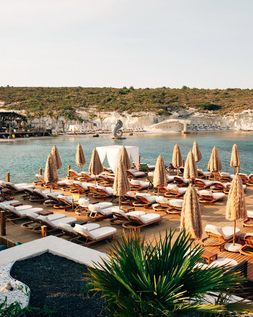 Çeşme'nin popüler happy hour ve gece mekanlarından biri olan Isla Alaçatı'da, denizin üzerinde uzanan geniş ahşap iskelede sıra sıra dizilmiş beyaz minderli konforlu şezlonglar ve doğal hasır şemsiyeler, berrak turkuaz sularla çevrili koyda yeşil tepelerin ve uzaktaki etkileyici denizatı heykelinin eşlik ettiği bir atmosfer sunuyor.