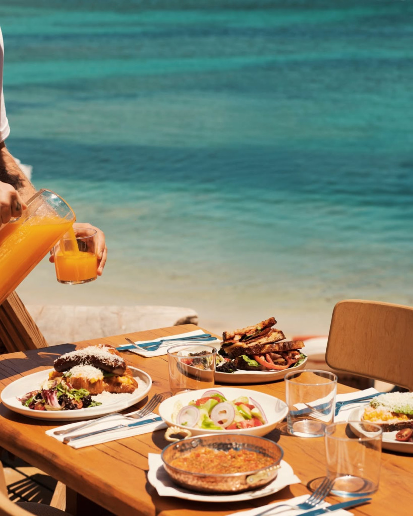 Çeşme'deki en iyi happy hour ve gece mekanlarından birinde, turkuaz denizin kıyısında güneşli bir günde, bir kişinin sürahiden portakal suyu doldurduğu, üzerinde zengin kahvaltı tabağı, salata, sandviçler ve menemenin yer aldığı ahşap bir masanın nefis bir öğünü gösteren canlı kare.