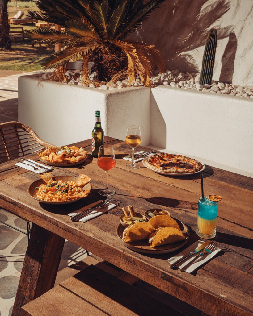 Cunda'nın güneşi altında, rustik ahşap bir masada sunulan pizza, makarna, çıtır deniz ürünleri, takolar ve renkli kokteyller, adanın zengin gastronomik deneyimini ve Ege'nin taze lezzetlerini gözler önüne seriyor.