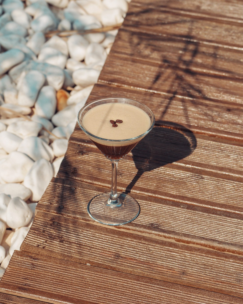 Cunda Seyahat Rehberi'nden ilhamla, güneşin parladığı ahşap bir iskelede, üzerindeki üç kahve çekirdeğiyle süslenmiş kremalı köpüklü bir espresso martini, beyaz çakıl taşlarının dingin fonunda Cunda'nın zarif ve keyifli molalarını vurguluyor.