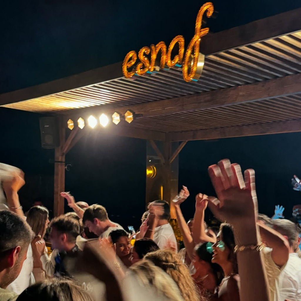 Gece vakti, parlak sarı 'esnaf' tabelasının aydınlattığı bir alanda, Çeşme'nin hareketli gece mekanlarından birinde ellerini havaya kaldırmış, müzik eşliğinde coşkuyla dans eden gençlerin oluşturduğu kalabalık bir eğlence sahnesi.