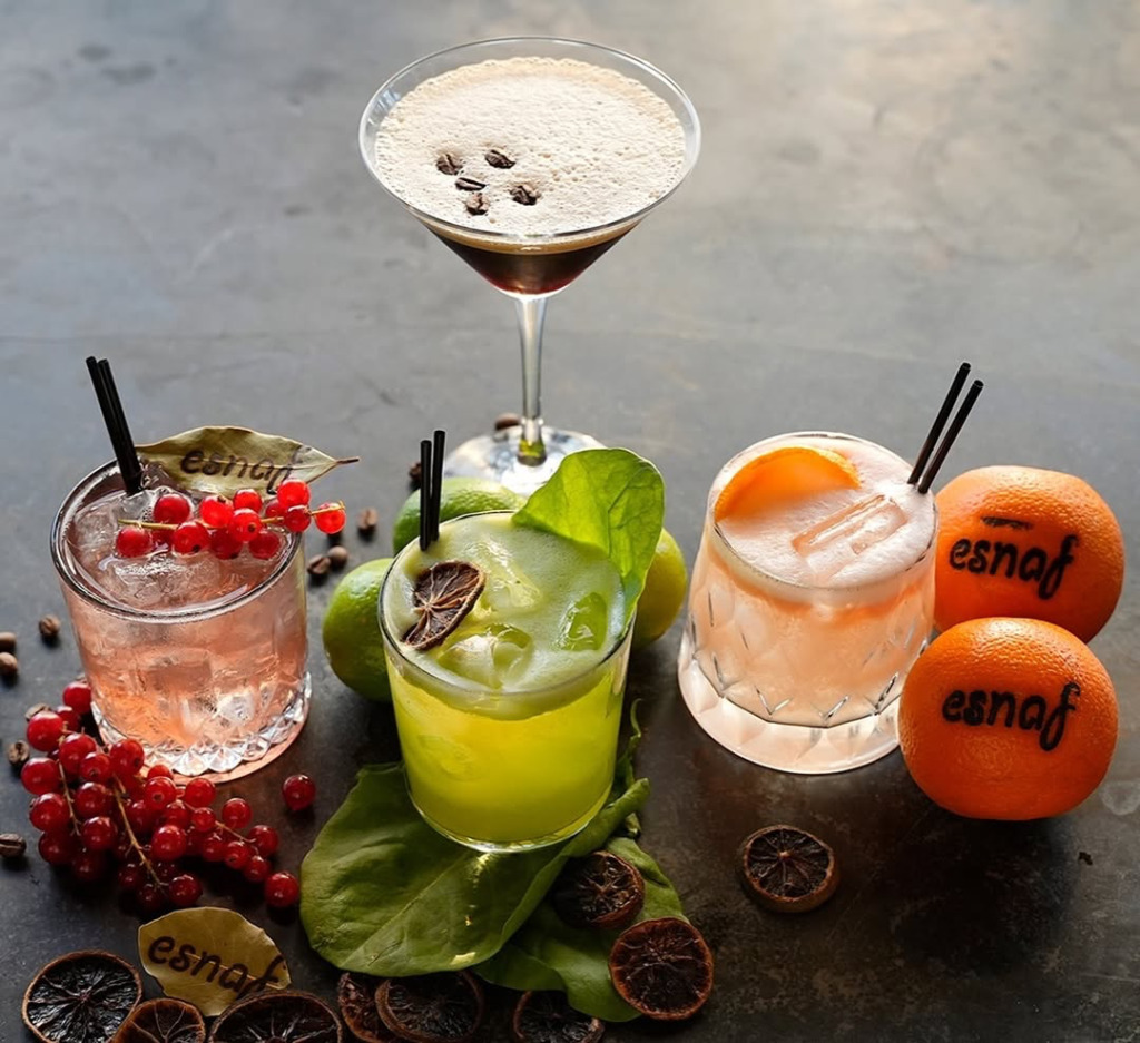 Çeşme'nin hareketli happy hour ve gece mekanları atmosferini yansıtan bu görselde, üzerinde Esnaf markasının belirgin olduğu frenk üzümlü, narenciyeli ve köpüklü espresso martiniler gibi renkli kokteyller, taze meyve ve kahve çekirdekleriyle özenle süslenmiş olarak masada iştah açıcı bir şekilde duruyor.