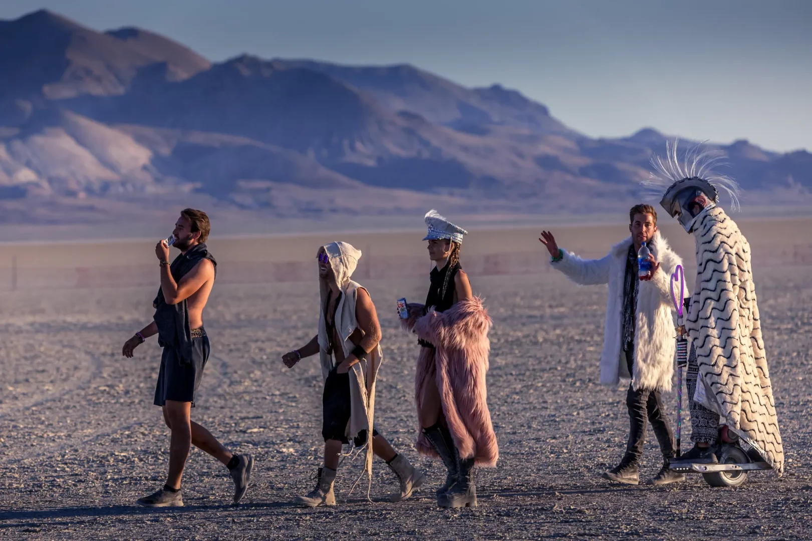 Geniş ve tozlu bir çöl arazisinde, Burning Man festivalinin ruhunu yansıtan çeşitli ve özgün kostümler giymiş beş katılımcı, dağlık bir fonun önünde yürüyor, en sağdaki kişi tek tekerlekli bir araç üzerinde ilerliyor.