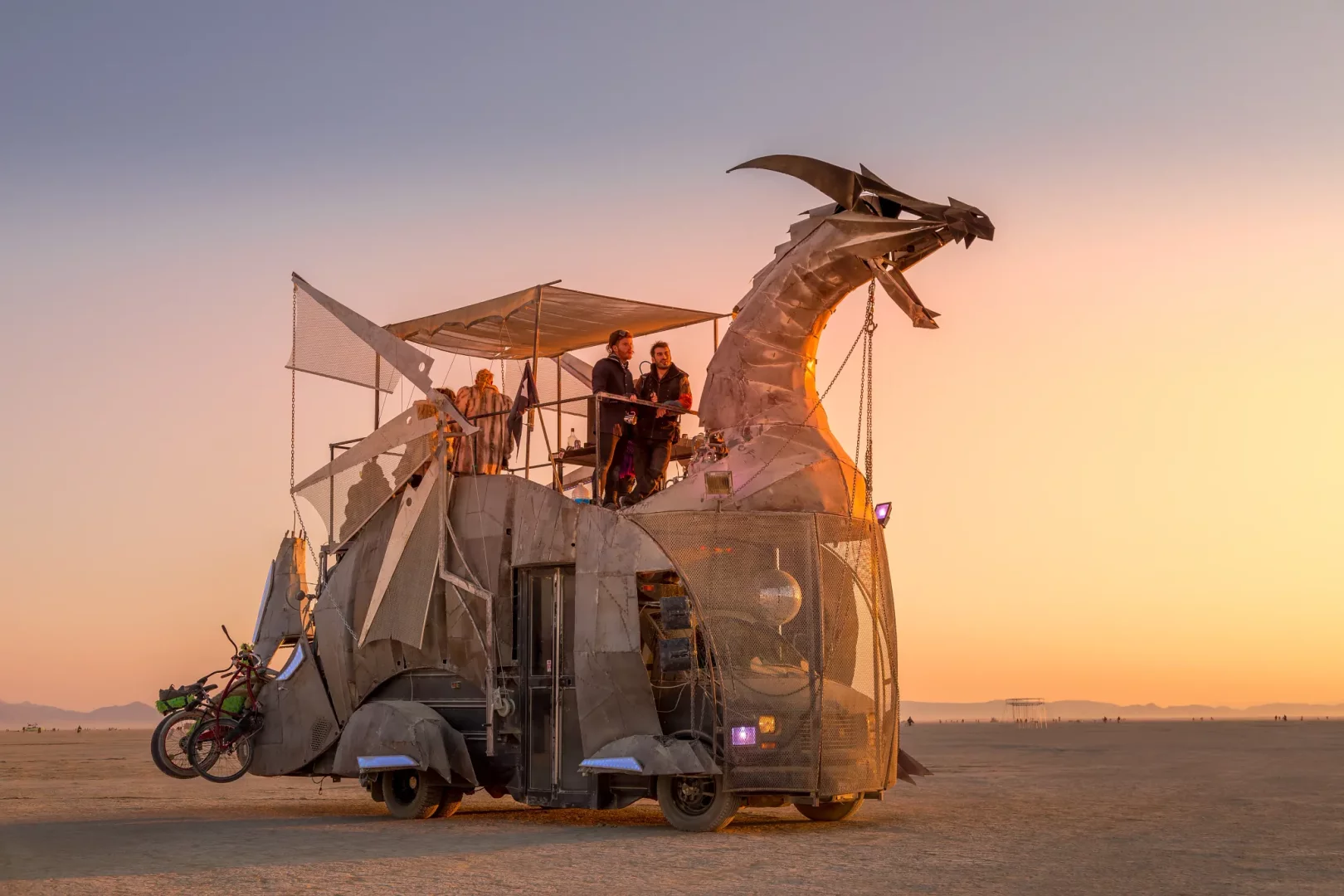 Gün batımının kızıl ve turuncu tonlarıyla aydınlanan uçsuz bucaksız kurak bir göl yatağında, metalik ejderha başlı ve katlı yapısıyla Burning Man festivaline özgü görkemli bir sanat aracı, üst güvertesindeki birkaç kişiyle birlikte duruyor.