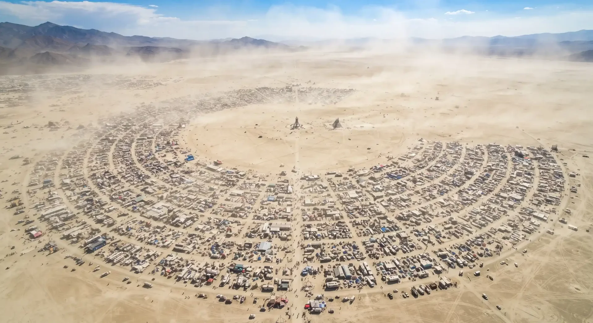 Havadan çekilmiş geniş açılı bir görüntüde, Nevada çölünün tozlu ve açık arazisinde yarı dairesel, konsantrik bir düzende kurulmuş binlerce çadır, karavan ve geçici yapıdan oluşan Burning Man festival alanı görülüyor; merkezde ise festivalin ikonik figürü The Man ve The Temple belirgin bir şekilde duruyor.