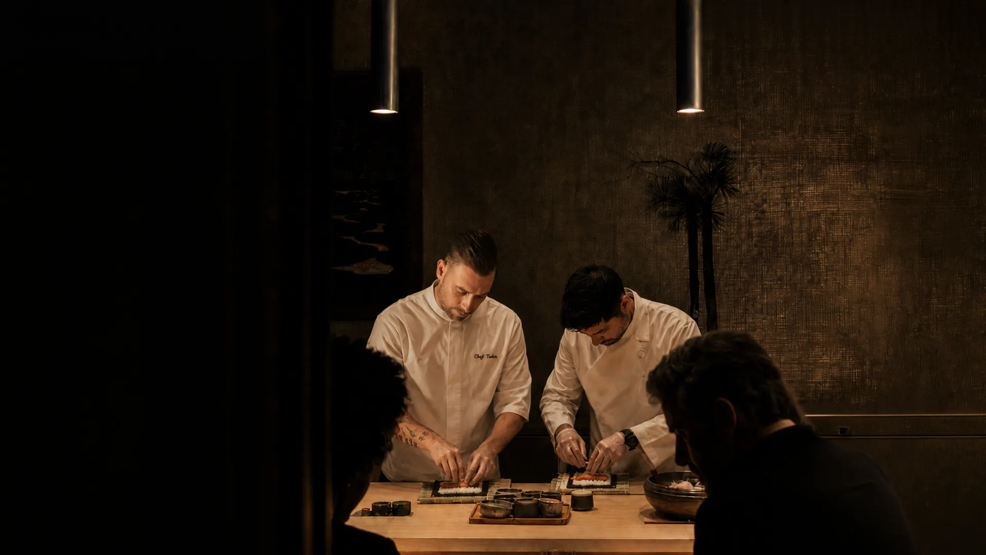 Elite World İstanbul Taksim'de düzenlenen Sushi Workshop'ta, beyaz şef üniformalı Chef Tin Tin ve Mert Karakuş'un katılımcılar önünde Japon mutfağının temel tekniklerini ustalıkla sergileyerek sushi hazırladığı, sıcak ışıklandırmalı bir an.