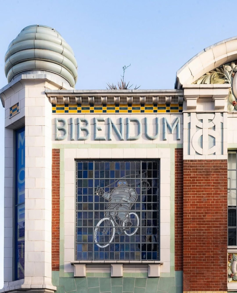 Açık mavi bir gökyüzünün altında, üzerinde BIBENDUM yazısı bulunan Art Nouveau mimarisine sahip bir binanın cephesinde, Michelin Adamı'nın bisiklet sürerken tasvir edildiği renkli bir vitray pencere dikkat çekiyor.