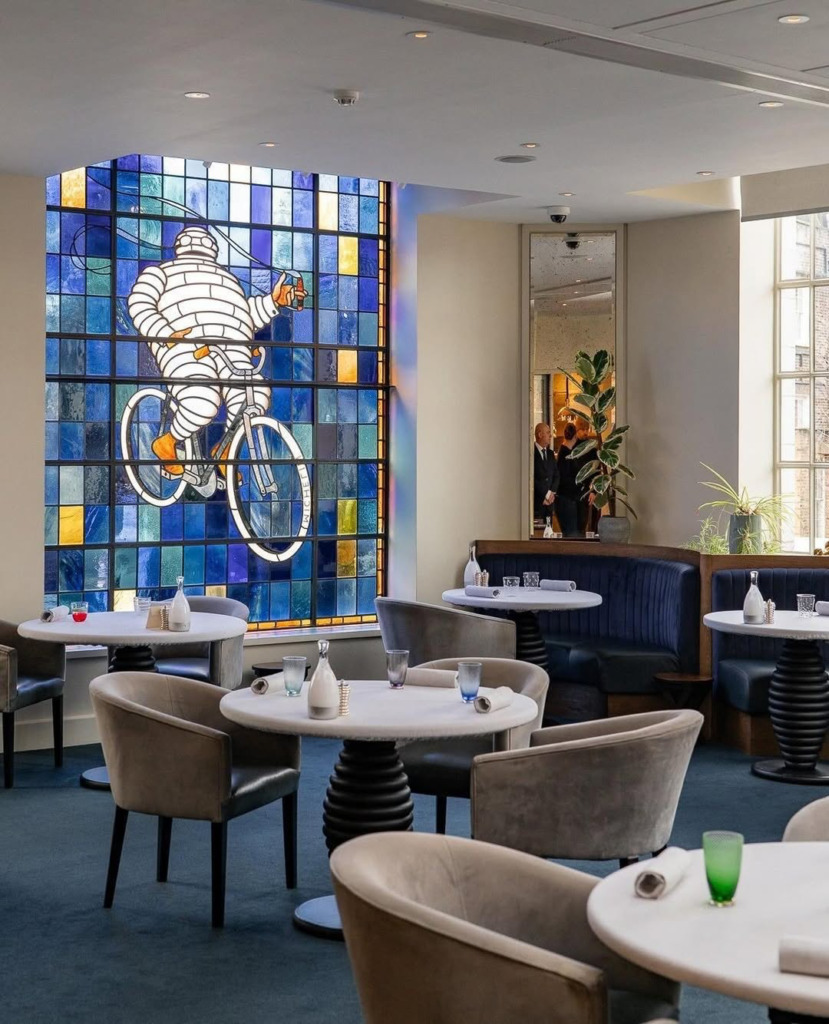Mavi tonlarında karolarla süslenmiş büyük bir vitray pencerenin önünde yer alan şık bir restoranın iç görünümü; pencerede bisiklete binen ve bir kadeh tutan ikonik Michelin Adamı figürü betimlenirken, mekanda mermer tablalı yuvarlak masalar, kadife kaplı sandalyeler ve koyu mavi halı döşemeler dikkat çekiyor.