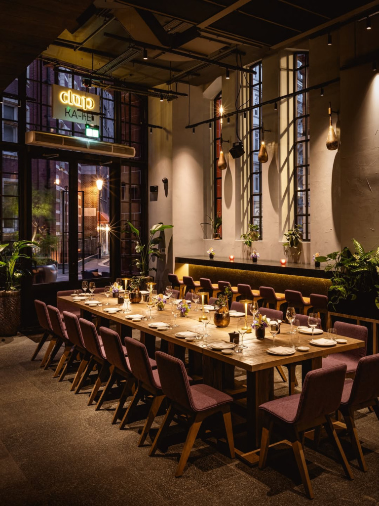 Modern ve şık bir restoranın loş ışıklı iç mekanını gösteren bu fotoğrafta, mor sandalyelerle çevrili, tabaklar ve kadehlerle özenle hazırlanmış uzun bir ahşap masa dikkat çekiyor; arka planda ise yeşil bitkilerle süslü bir bar alanı, büyük pencerelerden görünen akşam caddesi manzarası ve üstte parlayan CLUP KA-HE neon tabelası mekana sıcak ve davetkar bir atmosfer katıyor.