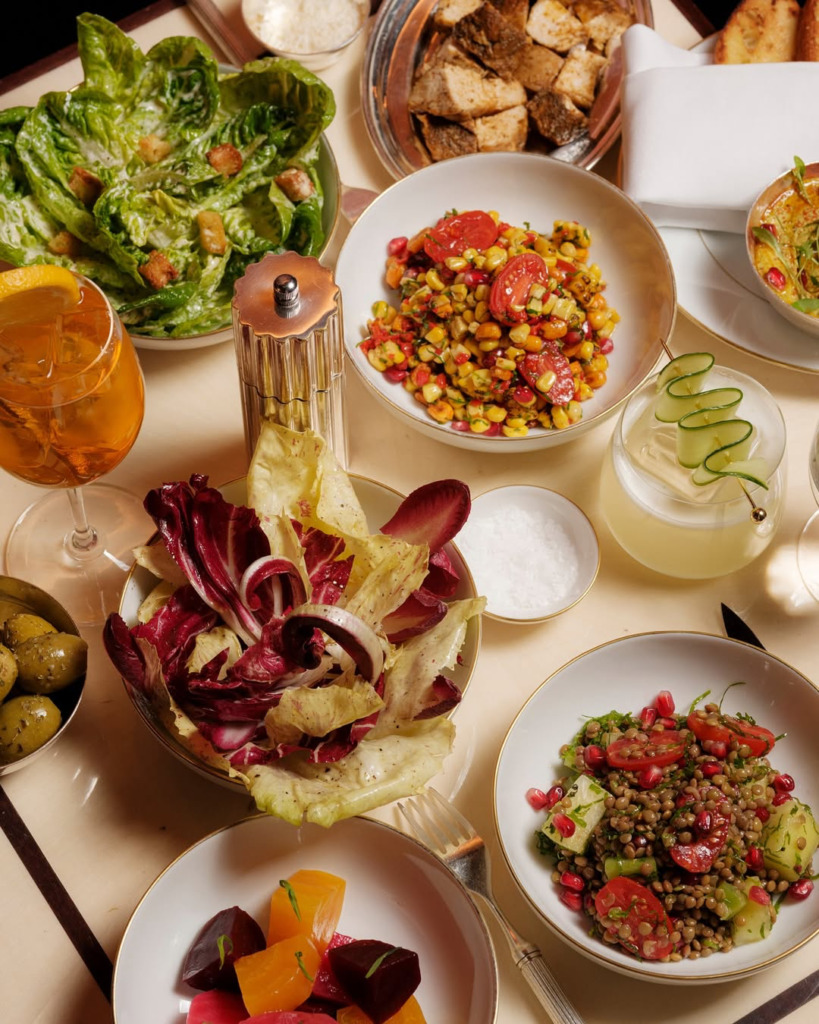 Bir masanın üzerinde, taze yeşilliklerden oluşan bir Sezar salatası, mısır, domates ve nar taneleri içeren renkli bir mısır salatası, kırmızı ve açık renkli marullardan oluşan bir radicchio salatası, küp kesilmiş pancar parçacıkları ve mercimek salatası gibi çeşitli lezzetli yemekler ve aperatif içecekler şık beyaz kaselerde servis edilmiş durumda.