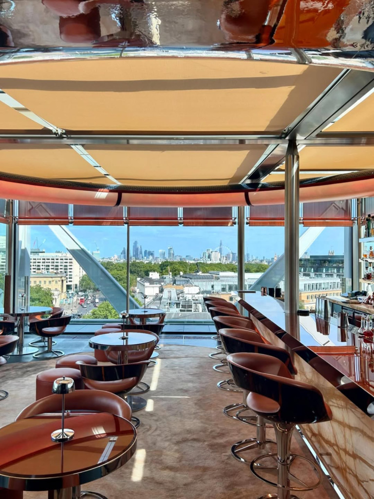 Şehir manzarasına bakan geniş pencereleri olan modern bir çatı katı barının iç görünümü, kahverengi deri sandalyelerle çevrili parlak yüzeyli yuvarlak masalar ve uzun bar tezgahının önünde sıralanmış yüksek taburelerle şık bir ortam sunuyor.