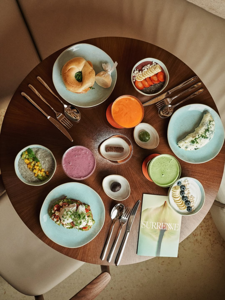 Üstten çekilen bu karede, yuvarlak ahşap bir masanın üzerinde özenle dizilmiş simit, çilek ve muz dilimli acai kasesi, taze sıkılmış portakal suyu, yeşillikli omlet, mangolu chia pudingi, mor ve yeşil smoothieler, granola ve yaban mersinli yoğurt kasesi gibi çeşitli sağlıklı ve besleyici kahvaltı seçenekleri ile 'SURRENNE' başlıklı bir dergi göz alıcı bir düzenlemeyle sunuluyor.