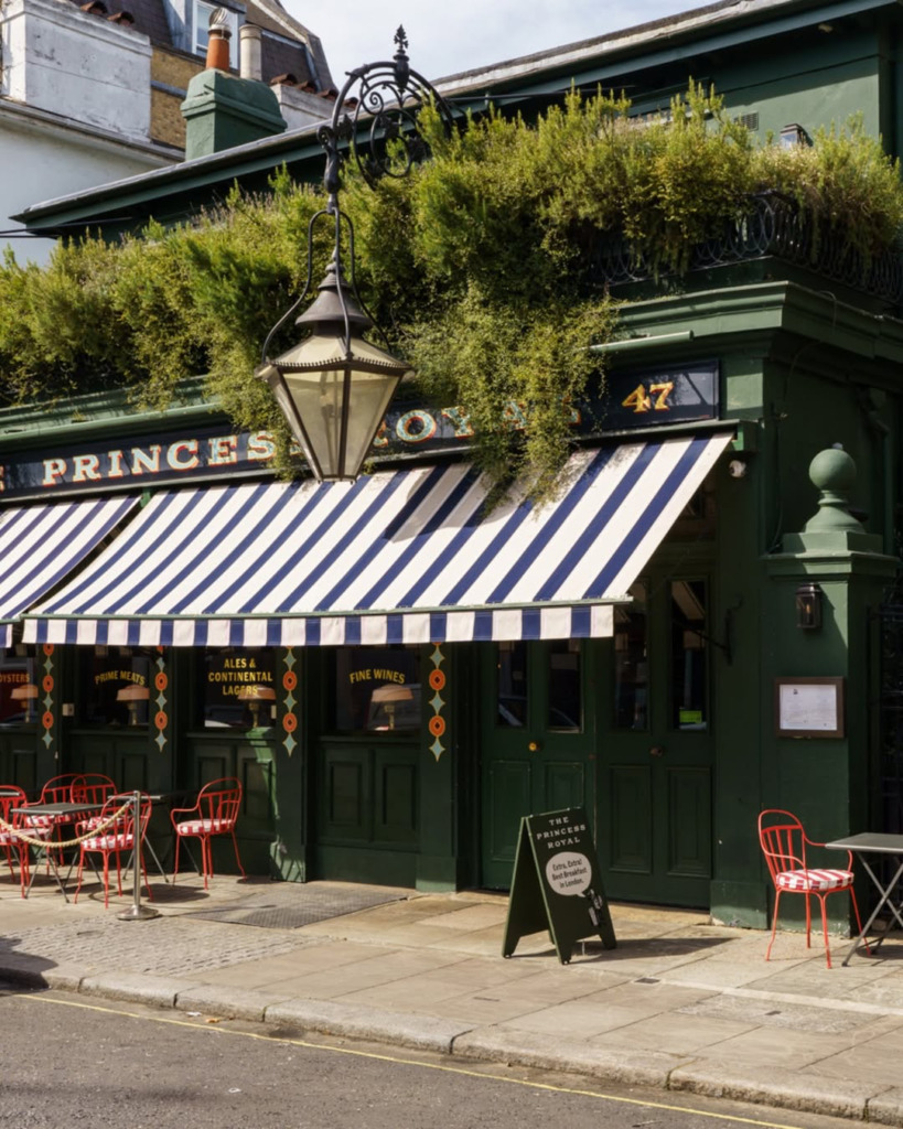 The Princess Royal adlı koyu yeşil bir pub ve restoranın dışarıdan çekilmiş görüntüsünde, mavi beyaz çizgili tenteler, kaldırımda kırmızı metal sandalyeler ve masalar, ön cephede asılı dekoratif bir fener ve çatıda bol yeşillik dikkat çekmektedir.