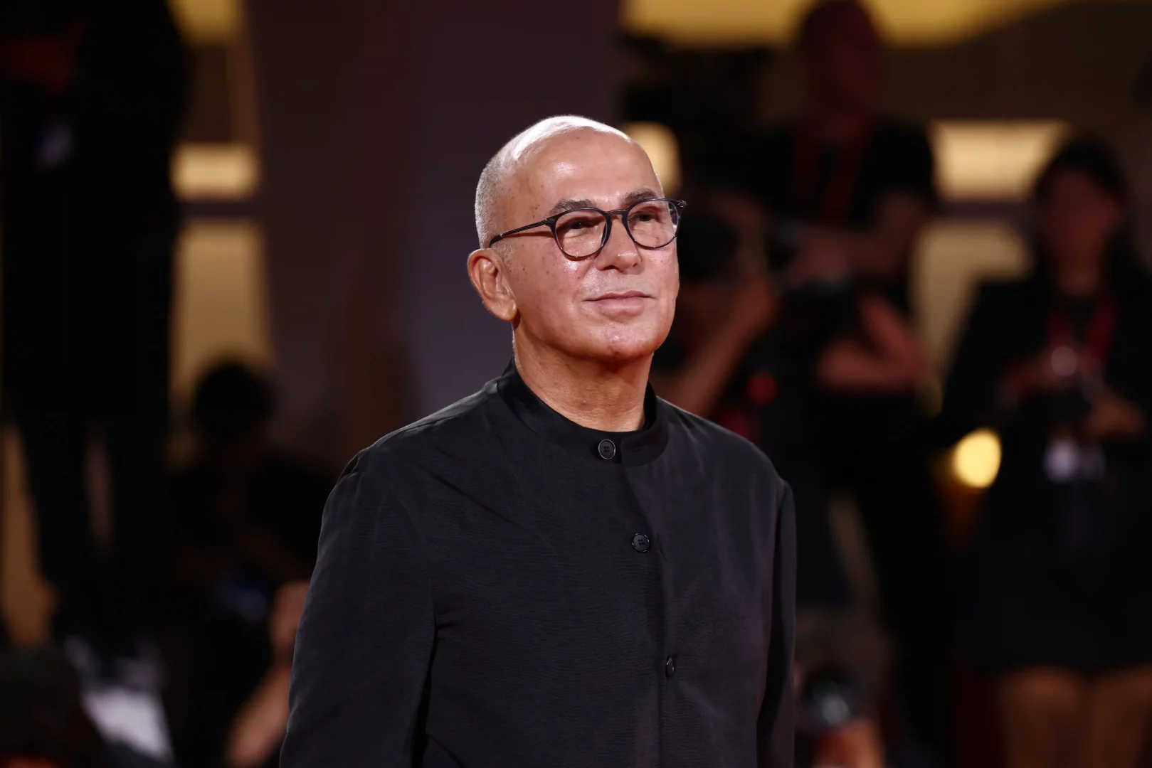 Giorgio Armani'nin hayatını konu alacak televizyon dizisinin yönetmen koltuğunda oturacak Ferzan Özpetek, kel kafalı, modern yuvarlak gözlüklü ve siyah yakasız gömleğiyle, bir etkinliğin karanlık fonunda hafifçe yukarıya bakarak zarifçe gülümsüyor.