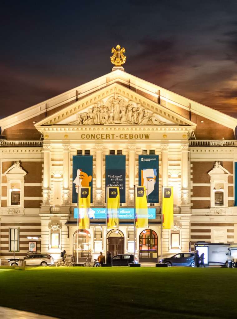 Akşam karanlığında, önünde yeşil bir çim alan ve birkaç araç bulunan, Concertgebouw yazılı klasik tarzda aydınlatılmış bir binanın cephesinde, Amsterdam Dance Event logosunu ve etkinlik yazılarını taşıyan uzun sarı ve turkuaz renkli afişler, Amsterdam'ın sanat ve kültür dolu ruhunu yansıtıyor.