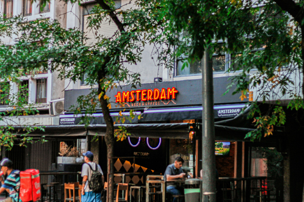 Akşamüstü bir Amsterdam sokağında, parlak neon tabelasıyla dikkat çeken Amsterdam Beer & Coffee barının önünde bisikletli bir kurye ve yoldan geçenler görülürken, bir müşteri masada telefonuna bakarak şehrin canlı gece hayatı enerjisini deneyimliyor.