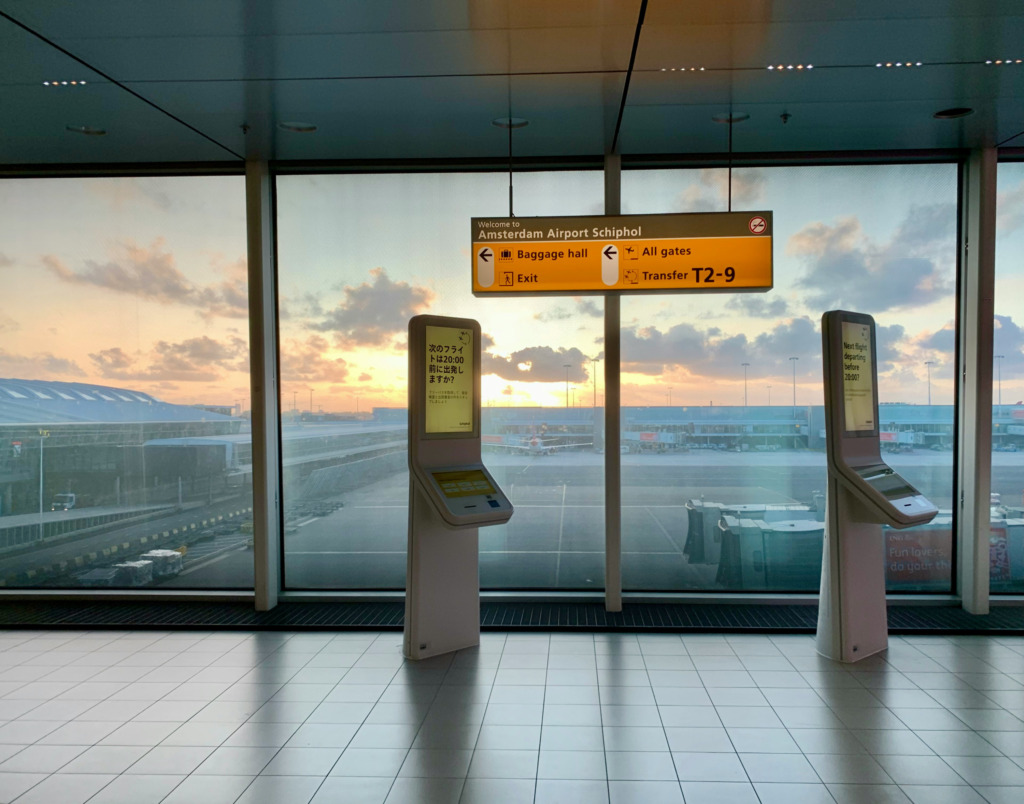 Amsterdam Schiphol Havalimanı terminalinden çekilen bu geniş açılı görselde, gün batımının turuncu ve mor tonlarına bürünmüş gökyüzünün altında, apron alanında bekleyen uçaklar ve yolculara yön gösteren Amsterdam Havalimanı tabelası ile dijital bilgilendirme ekranları belirgin bir şekilde görülüyor, bu da Amsterdam seyahati için havayolu ulaşımının önemini vurguluyor.