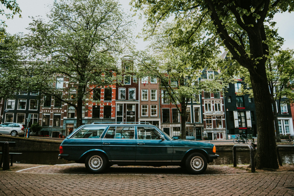 Amsterdam kanallarının kenarında, koyu teal rengi nostaljik bir station wagon araba Arnavut kaldırımlı yolda park etmişken, arkada sıraya dizilmiş pastel tonlu tarihi evler ve gür yeşil ağaçlar, Amsterdam Seyahat Rehberi'nde bahsettiğimiz Kanallar şehrinin dinginliğini ve zamansız güzelliğini vurguluyor.