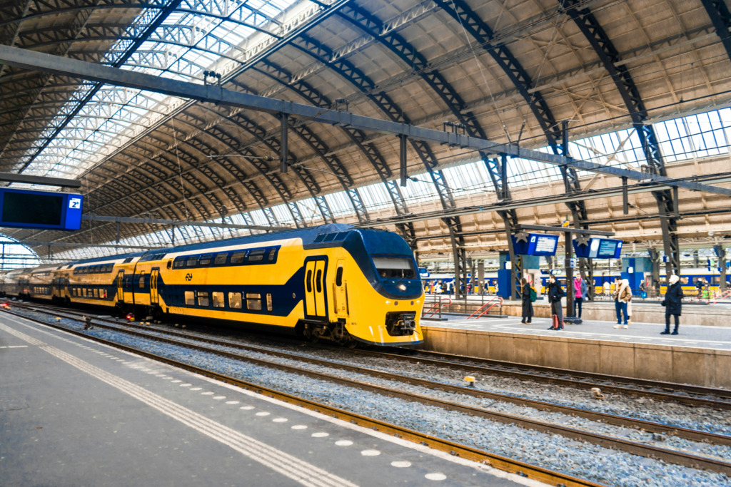 Geniş cam tavanlı modern bir tren istasyonunda sarı ve mavi renklerdeki bir yolcu treni peronda beklerken, Amsterdam seyahatleri için varış veya ayrılış yapan yolcuların olduğu hareketli ortam, demiryoluyla kolay ulaşımın pratik bir örneğini sunuyor.
