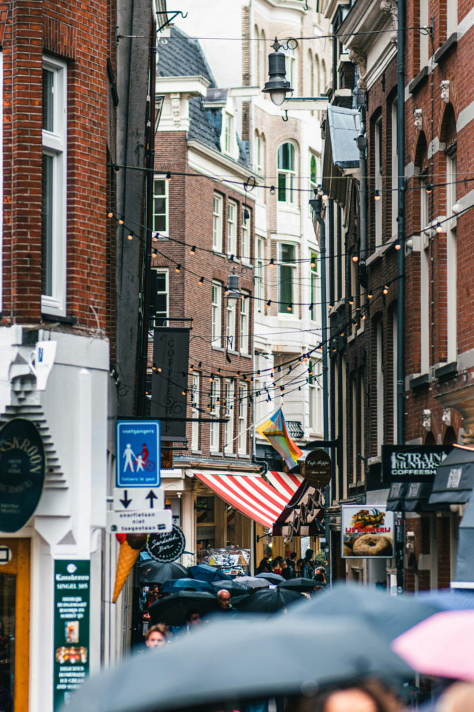 Yağmurlu bir günde Amsterdam'ın karakteristik dar sokaklarından birinde, tarihi tuğla binalar arasında asılı dekoratif ışıklandırmalar ve şemsiyelerle kaplı kalabalık insanlar, şehrin canlı alışveriş atmosferini ve geleneksel dükkanlarını yansıtıyor.