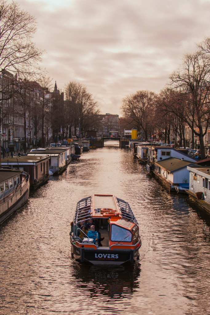 Amsterdam'ın UNESCO Dünya Mirası listesindeki büyüleyici kanallarından birinde, sonbahar veya kış mevsimini işaret eden yapraksız ağaçlar ve tarihi binaların sıralandığı kıyılar arasında, gün batımının sıcak yansımalarıyla ilerleyen bir tur teknesi, şehrin kanallar ile şekillenmiş eşsiz seyahat ruhunu gözler önüne seriyor.