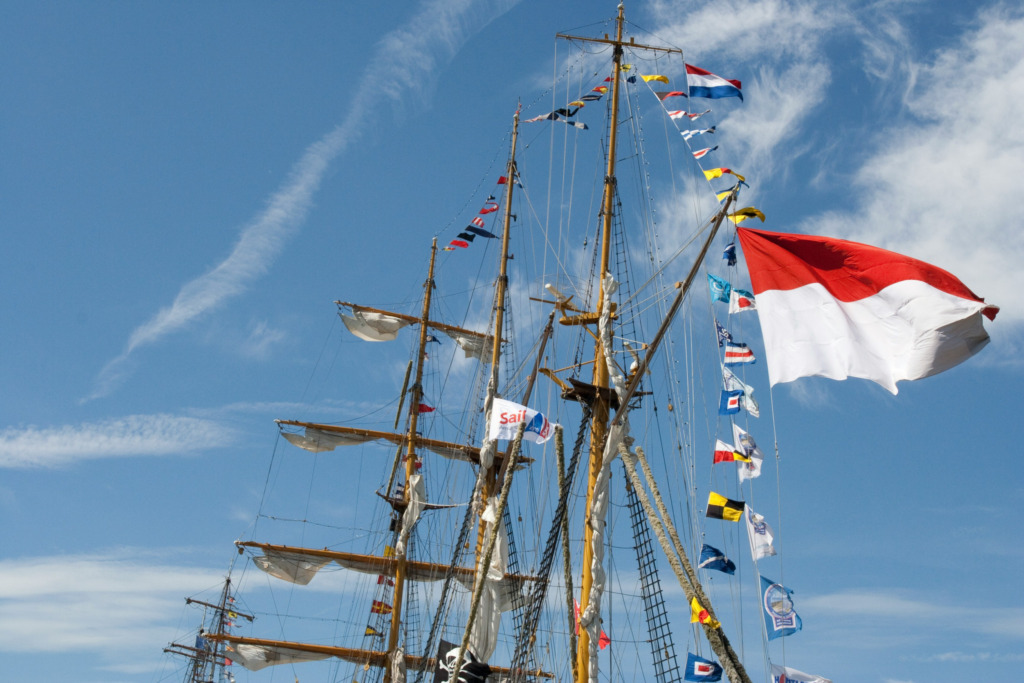Mavi gökyüzü altında, direkleri ve donanımı rengarenk bayraklarla süslenmiş büyük bir yelkenli gemi, Amsterdam'ın kanallarla iç içe denizcilik geleneğini ve şehirdeki yıl dönümü kutlamalarını akla getiren görkemli bir festival atmosferi sunuyor.