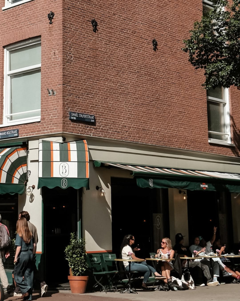 Amsterdam'ın hareketli De Pijp bölgesinde, Ferdinand Bolstraat ve Daniel Stalpertstraat'ın köşesindeki Bar Bellini'nin güneşli dış mekân masalarında, çeşitli insan grupları keyifli sohbetler eşliğinde içeceklerini yudumlayarak şehrin sosyal atmosferinin tadını çıkarıyor.