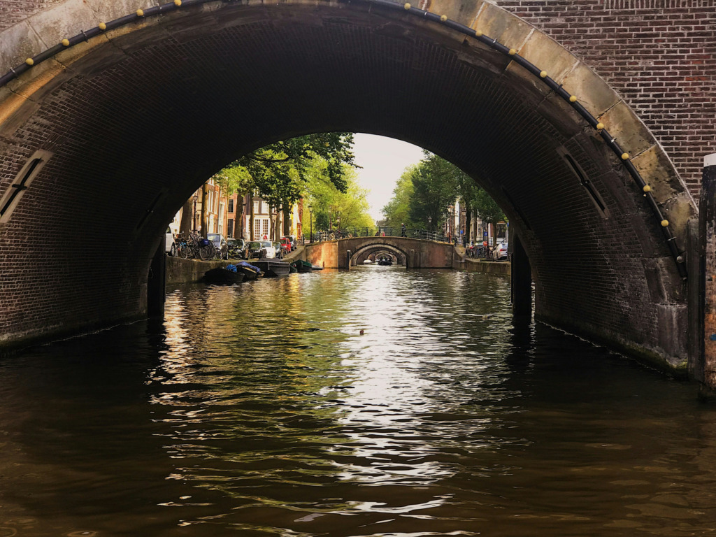 Büyük, tuğla kemerli bir köprünün altından bakıldığında, Amsterdam'ın UNESCO Dünya Mirası Listesi'ndeki Kanallar Bölgesi'nin karakteristik manzarası, tarihi evlerin sıralandığı yemyeşil ağaçlı kıyılar ve su üzerinde ilerleyen küçük teknelerle ışıl ışıl parlayan kanal, bu büyülü şehrin kalbini gözler önüne seriyor.