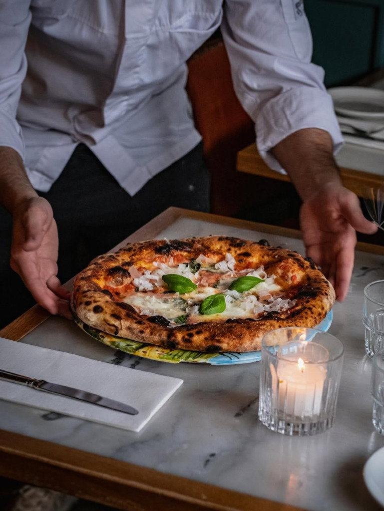 Amsterdam seyahat rehberi önerilerinden Ceppis İtalyan restoranında, beyaz şef ceketli bir aşçının, üzerinde mozzarella, baharatlı salam ve taze fesleğen bulunan, odun ateşinde pişmiş çıtır kabuklu pizzayı mermer bir masaya servis ettiği, yanan mum ışığıyla aydınlatılmış şık bir an.