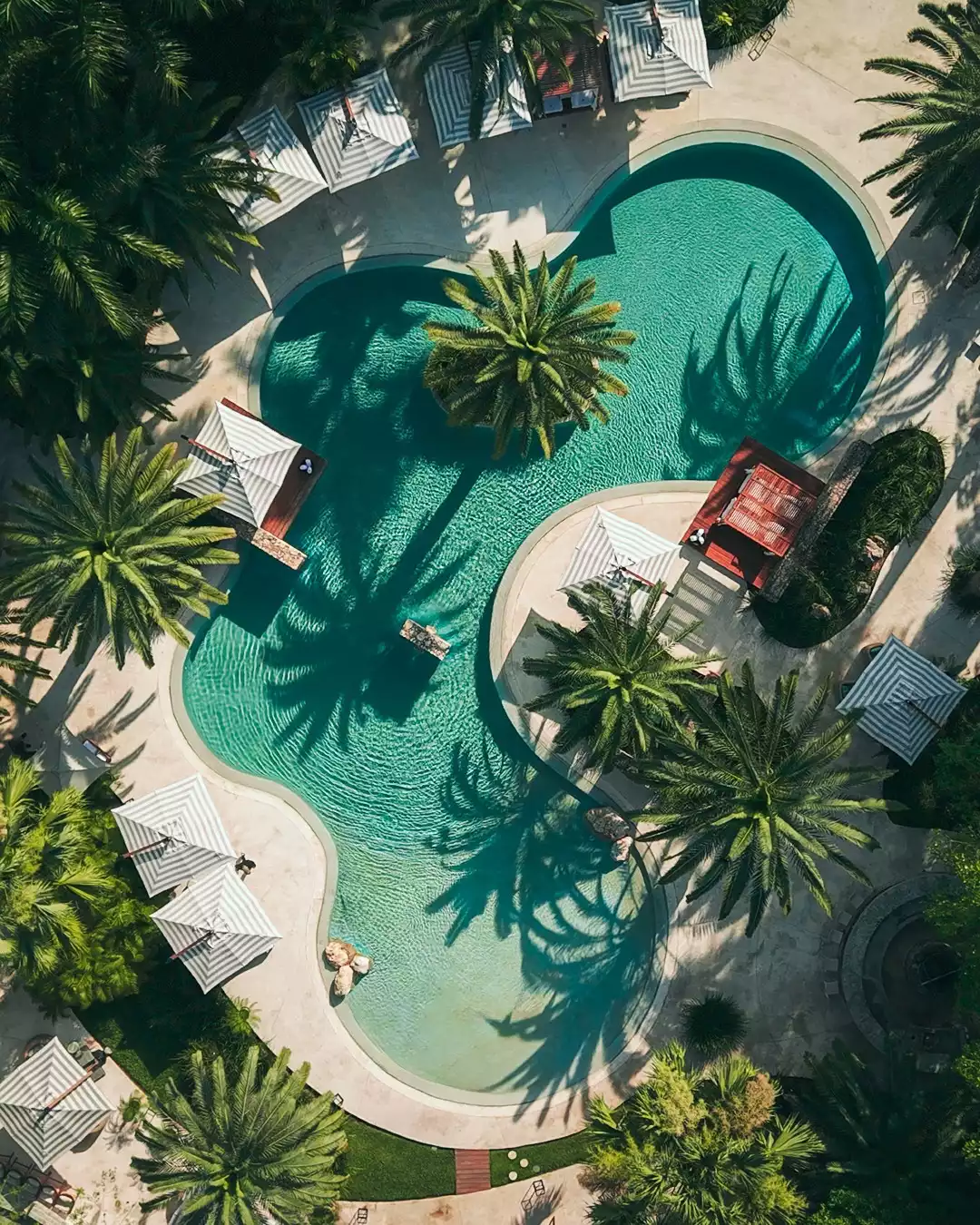 Chablé Yucatán gibi lüks bir tatil köyünde çekilmiş kuşbakışı bu fotoğrafta, organik formlu turkuaz rengi havuzun etrafında beyaz-mavi çizgili şemsiyeler ve palmiye ağaçlarının suya düşen uzun gölgeleriyle dinlendirici bir atmosfer göz alıcı biçimde sergileniyor.