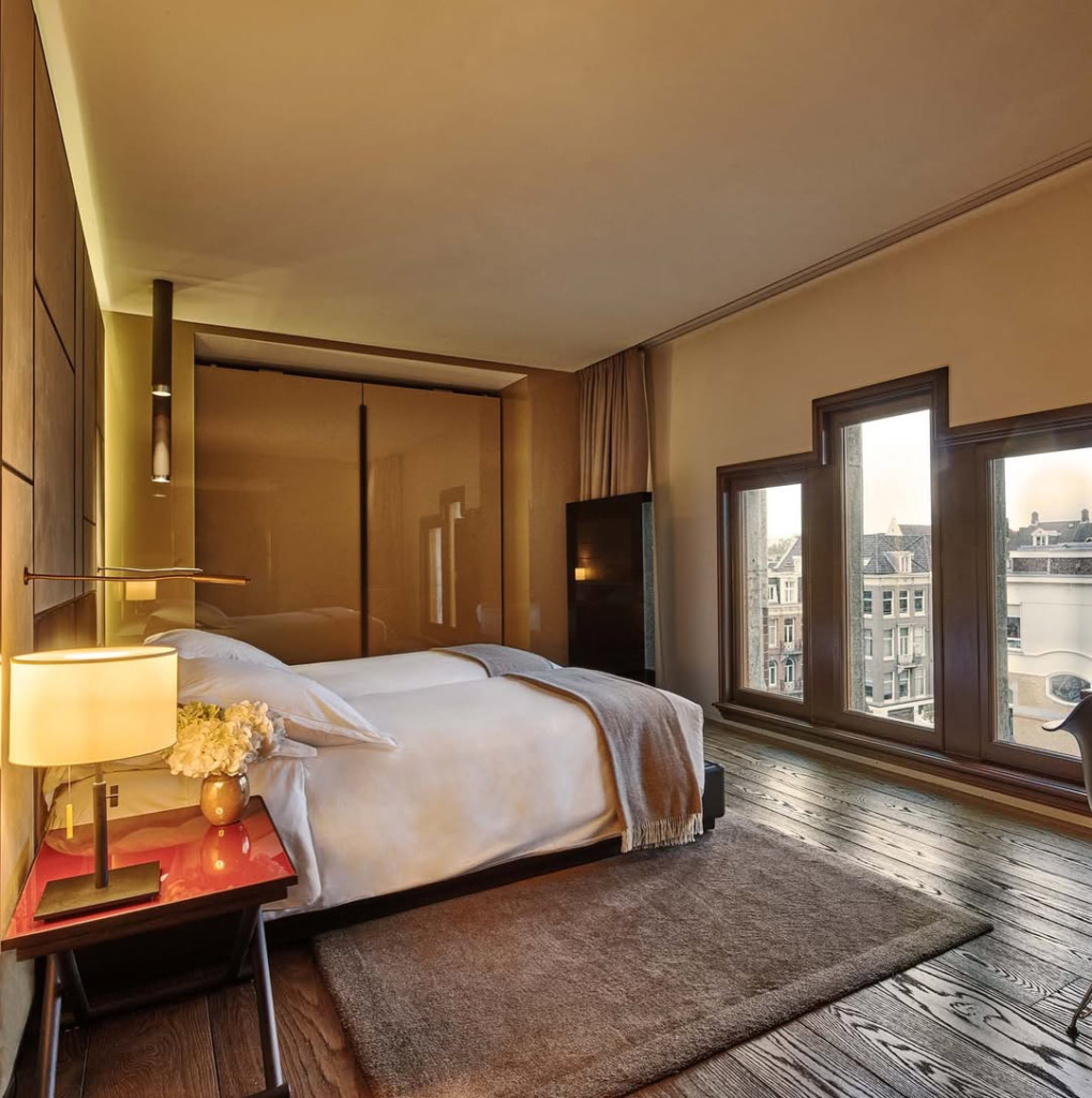 Conservatorium Hotel'in koyu ahşap parke zeminli, beyaz çarşaflı ve gri battaniyeli lüks yatak odası, kırmızı komodin üzerindeki zarif lambası ve penceresinden görünen tarihi Amsterdam şehir manzarasıyla huzurlu bir konaklama sunuyor.