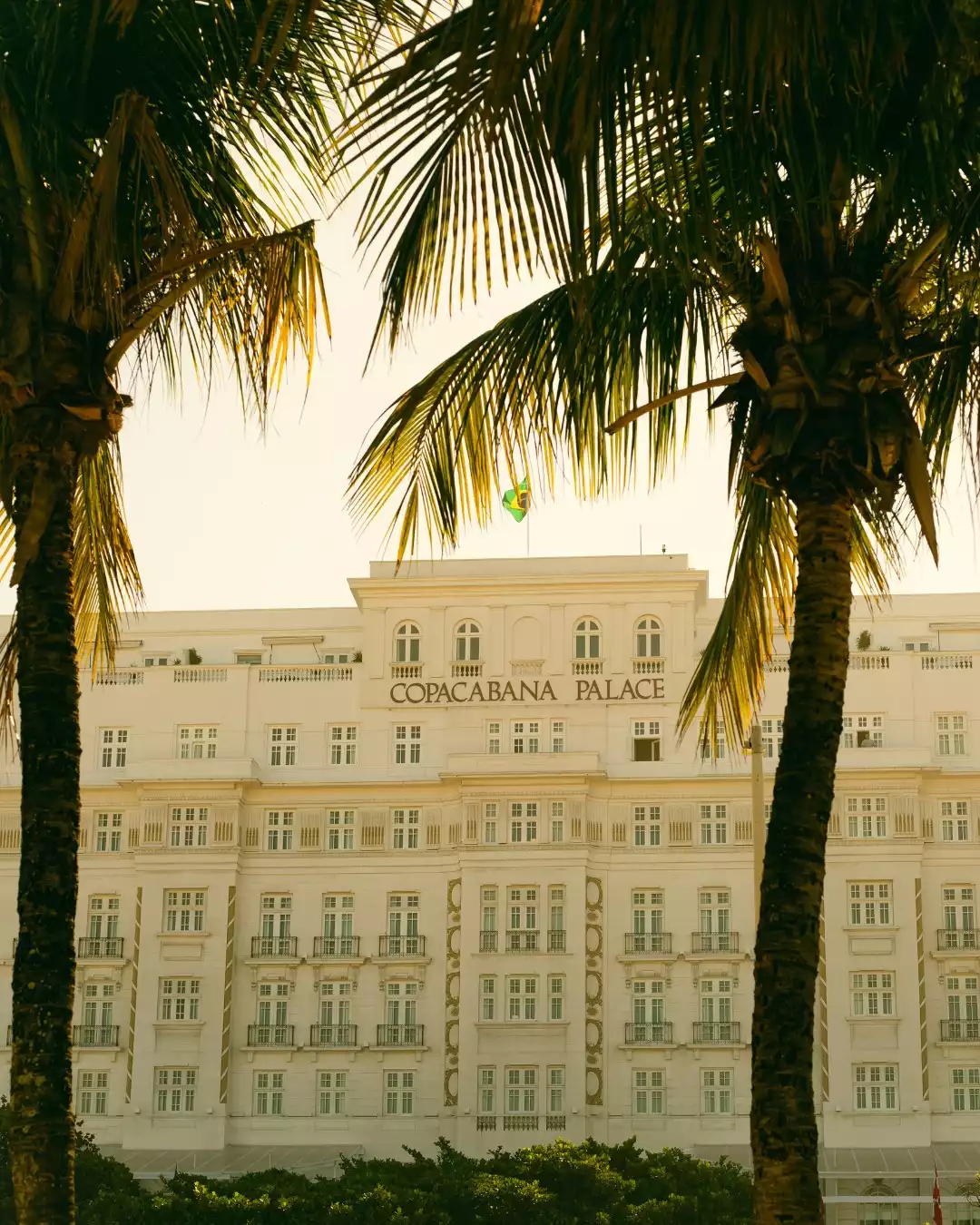 Cephesinde belirgin bir şekilde COPACABANA PALACE yazılı olan, iki yandan palmiye ağaçlarıyla çerçevelenmiş, çatısında Brezilya bayrağı dalgalanan, bembeyaz ve süslü lüks otel binası gün batımı ışığında parlıyor.