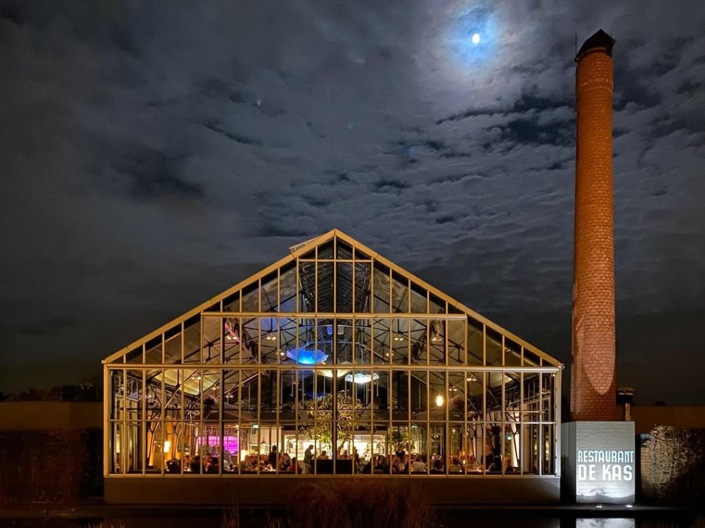 Ay ışıklı bulutlu bir gecede, Amsterdam'ın doğu yakasında, kendi bahçesinden taze ürünlerle ünlü De Kas restoranının 1926 yapımı cam serası, suya yansıyan sıcak ışıklarıyla aydınlanırken, yanında yükselen tuğla bacası dikkat çekiyor ve şehrin büyülü atmosferine eşsiz bir gastronomik deneyim katıyor.