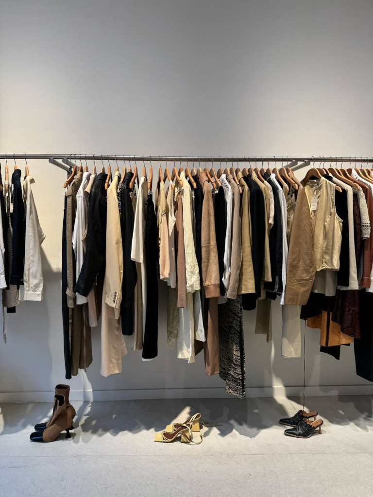 Amsterdam seyahat rehberi için önerilen Enso Vintage butiğinden, minimalist bir zarafetle küratörlüğü yapılmış raflarında asılı bej, siyah, krem ve kahverengi tonlarında çeşitli giysileri ve yerde sergilenen iki tonlu botlar, sarı topuklu sandaletler ile siyah desenli ayakkabıları gösteren mağaza içi görünüm, özel bir alışveriş deneyimini vurguluyor.